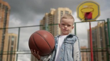 Kid boy is standing on basketball court holding ball and looking at camera. Child and sport concept. Outdoors activities in urban life for kids in residental district in city. Camera is around kid.