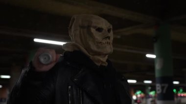 Halloween. Scary horror, an evil scary Scarecrow with bag on his head, in dark vestments and baseball bat on shoulder walk in an underground parking lot in night. Horror. Masked maniac at night