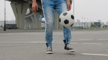 Footballer is exercising training outdoors juggling ball. Wearing casual style. Sports football skills concept. Man soccer player in jeans is training freestyle tricks with ball, legs close-up.