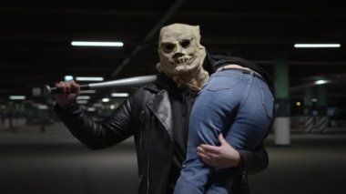 Homicidal maniac on Halloween or Doomsday. Male killer in scary scarecrow mask with baseball bat in his hands carries girl on victims shoulder in an underground parking lot at night