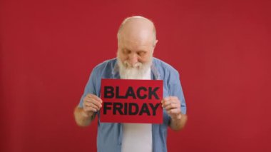 Big sale on Black Friday, big discount, Christmas sale. An elderly cheerful grandfather is dancing with a BLACK FRIDAY banner in his hands on a red background. High quality 4k footage