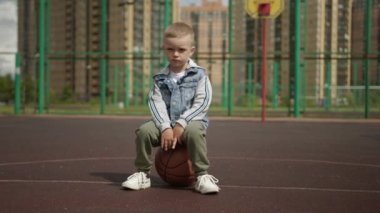 Child training and sport concept. Little boy in denim shirt is sitting on basketball ball on court in residental district in city and looking at camera. Outdoors activities in urban life for kids.
