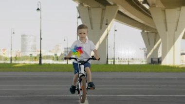 Child boy is riding a bike bicycle with pinwheel on asphalt road with cityscape background at summer day in slow motion. Modern urban life of kid. Outdoors activities for kids happy childhood concept.