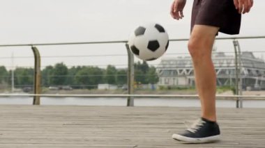 Sports football skills concept. Professional man footballer is juggling a ball with his feet on waterfront, legs close-up. Soccer player is training outdoors with ball improving skills in slow motion.