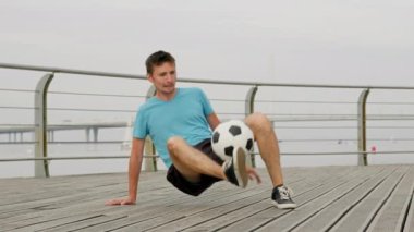 Soccer player is professionally juggling ball with feet sitting on waterfront in city. He performs difficult tricks in slow motion. Man footballer is training outdoors in freestyle. Sports concept.