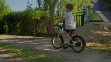 Child boy is riding a bike with windmill in the village on ground path road near the house at summer sunny warm day. Outdoors activities for kids and happy childhood concept. Kid on bicycle on nature.