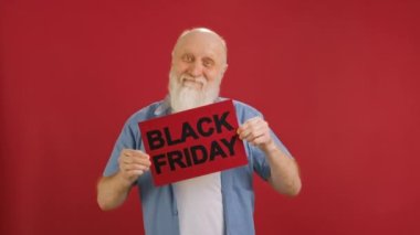 Elderly man with beard in white T-shirt and blue shirt raises banner with inscription BLACK FRIDAY, with place for advertising, rejoices at big sale or discount in store and dances on red background
