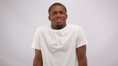 Portrait of young african american man wearing white t-shirt shrugging his shoulders and pointing with finger up saying no with serious face on white background. No gesture and disagree concept.