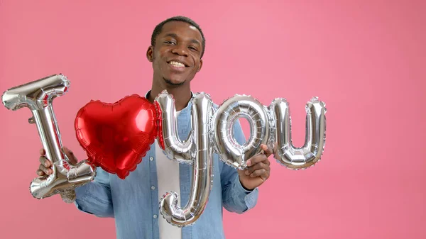 Smiling African-American teenager calls for love or confesses his love to loved one, holds word I love you from helium balloon in his hands, congratulates on Mother Day or Valentines Day