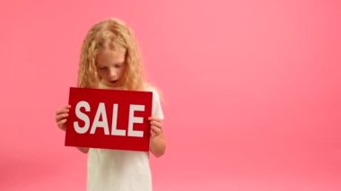 Cheerful blonde caucasian teenage girl in white T-shirt dances and smiles holding red banner with inscription sale on studio background. Girl advertises big sale on Black Friday and big discount