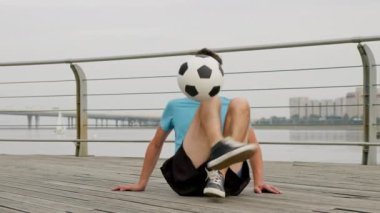 Professional footballer is juggling ball with feet sitting on waterfront in city. He performs difficult trick. Man soccer player is training outdoors in freestyle. Sports football skills concept.