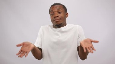 Confused african american man in white t-shirt shrugs shoulders in bewilderment, doubtful looks, spreads palms on white background. He doesnt know nothing. People and hesitation, doubting concept