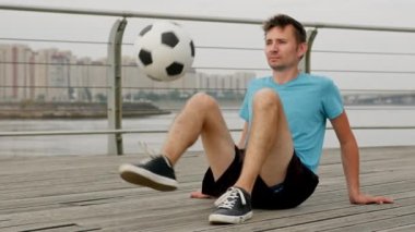 Young man soccer player is practicing kicks with ball sitting on waterfront honing his technical skills. Professional footballer is training, warming-up outdoors. Sports and football skills concept.