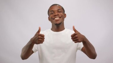 Smiling happy american african man wearing white t-shirt is showing thumbs up on white background looking at camera. Cheerful face expression and winner positive hands gestures of young guy.