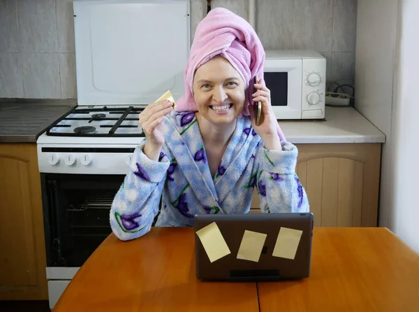 woman in bath towel on her head and wearing bathrobe working with laptop and documents in kitchen at home, woman accountant conducts an audit, home office concept