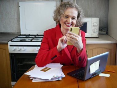Mutfakta dizüstü bilgisayarı ve banka kartıyla çalışan ceketli orta yaşlı bir kadın. Ev ofis konsepti, kapalı alan.