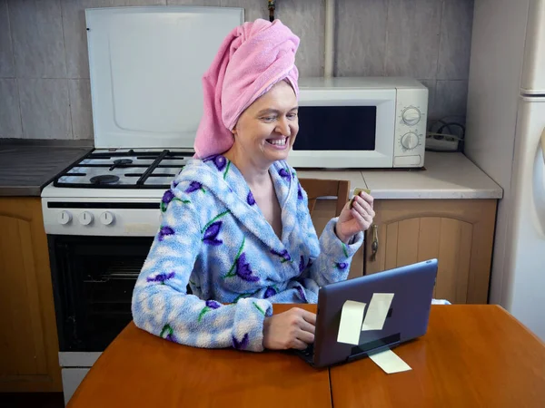 woman in bath towel on her head and wearing bathrobe working with laptop and documents in kitchen at home, woman accountant conducts an audit, home office concept