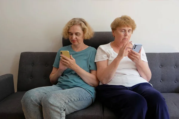 two women sitting on sofa at home and separately looking at their phones, loosing communication. People using their phones and sending texts as they sitting beside each other. Addiction to gadgets. Online vs offline concept
