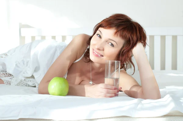 Aantrekkelijke jonge vrouw met een groene appel en glas water op bed — Stockfoto