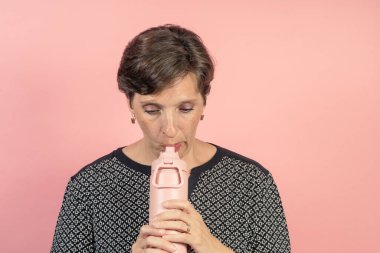 Brunette woman in her 50s in the studio with a pink background about to drink from a flask