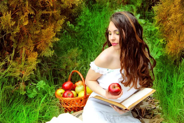 Mujer joven con manzana y libro en el huerto. Cesta de manzanas —  Fotos de Stock