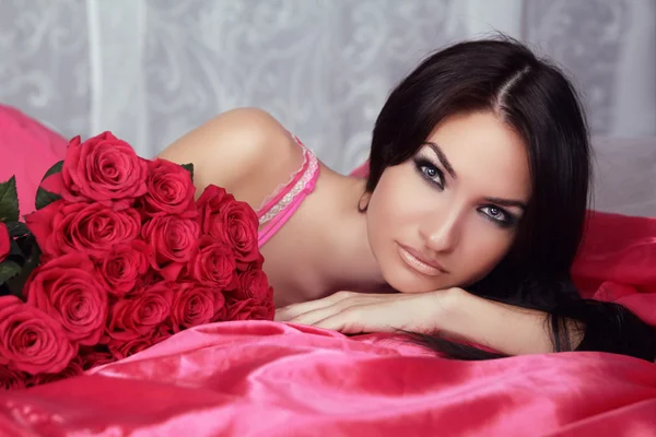 Retrato de belleza de chica morena con rosas rojas acostada en la cama — Foto de Stock