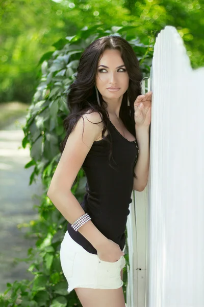 Attractive brunette woman posing near white fence at spring park — Stock Photo, Image