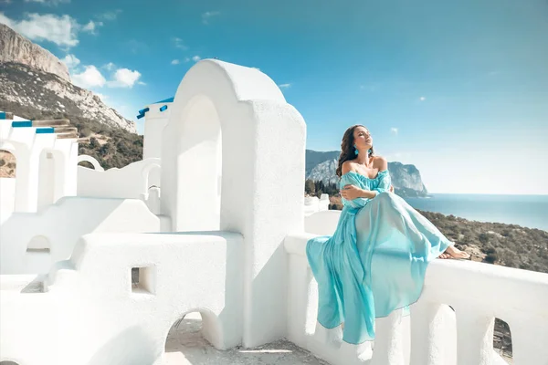 青い空の上にアーチによって白いバルコニーに座って青いシフォンドレスで巻き毛のロマンチックな女の子 サントリーニ島ギリシャを楽しむ美しい花嫁モデル — ストック写真