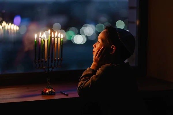 子供は窓辺のハヌッカのメノアに火をつけます 窓のそばに腰を下ろしていたキッパイの少年 ユダヤ人の祝日 伝統は宗教儀式です 最初の星だ ユダヤ教 — ストック写真