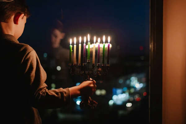 子供は窓辺のハヌッカのメノアに火をつけます 窓のそばに腰を下ろしていたキッパイの少年 ユダヤ人の祝日 伝統は宗教儀式です 最初の星だ ユダヤ教 — ストック写真