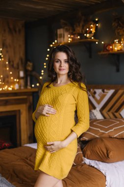 Sarı örgü elbiseli güzel hamile kadın karnına sarılıyor. Sonbaharda güzel bir hamilelik. Şükran Günü havası. Sıcak renkli yatak odasının içi sıcacık. Sonbahar Merhaba Ekim.