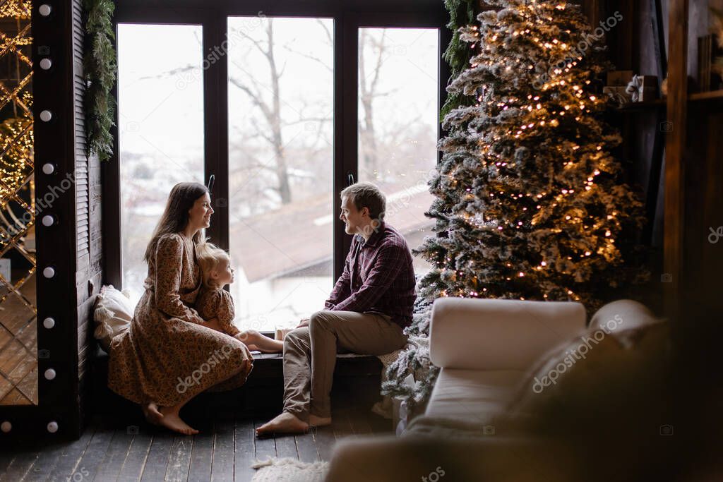 A full family celebrates Christmas at home on the windowsill. Nice and cozy loft interior with large windows. Happy parenting. Christmas spirit. Warm holiday atmosphere. Togetherness in new year