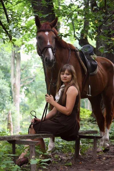 Young girl sitting on the bench and chestnut horse standing near — Stock Photo, Image