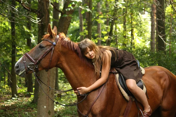 Tjej i klänning och bruna hästen stående i skogen — Stockfoto