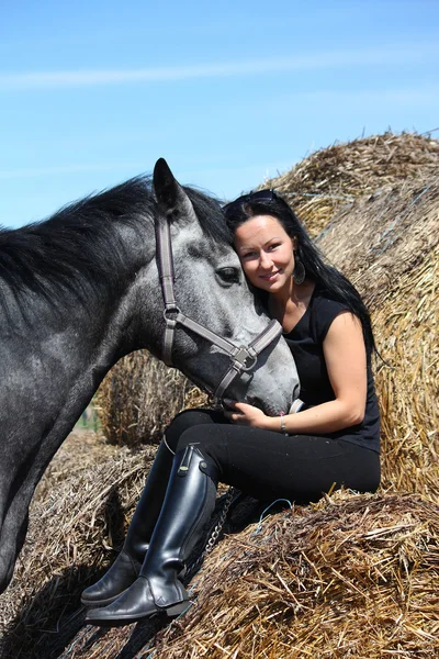 Beautiful woman sitting on hay bale and gray horse — Stock Photo, Image