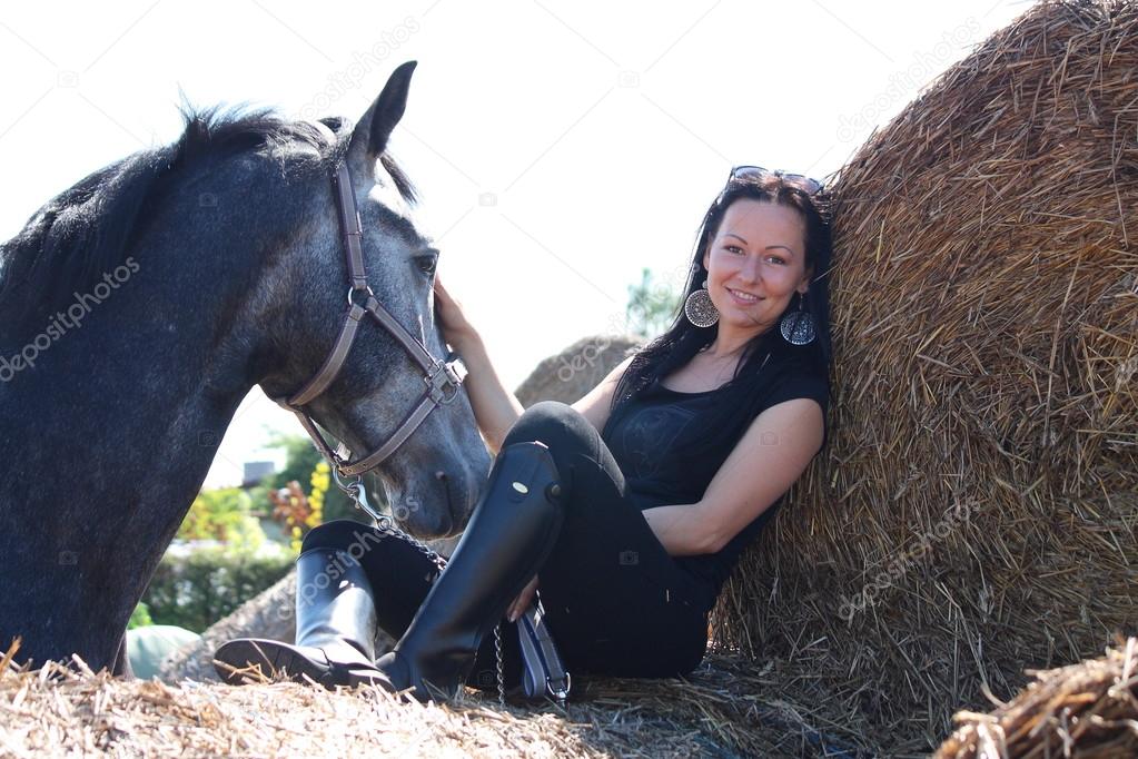 Beautiful woman sitting on hay bale and gray horse