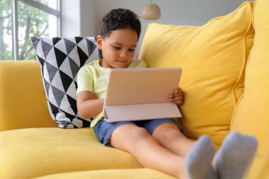 Tablet bilgisayarlı küçük Afro-Amerikalı çocuk evdeki kanepede çizgi film izliyor.
