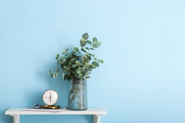 Renk duvarının yanındaki masada okaliptüslü saat ve vazo.