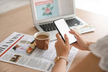 Masada kahve, gazete ve dizüstü bilgisayarlı cep telefonu kullanan bir kadın.