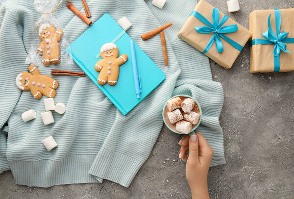 Female hand with cup of cacao, cookies, gifts, notebook and sweater on grunge background