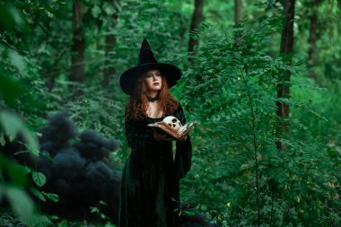 Yeşil ormanda büyü kitabı ve kafatası olan genç cadı.
