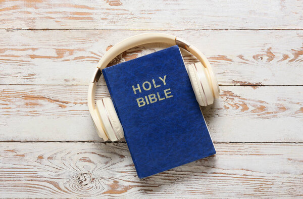 Holy Bible and modern headphones on light wooden background