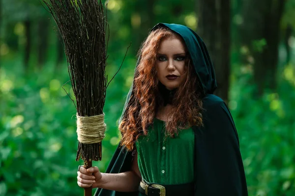 Young witch with broom in green forest