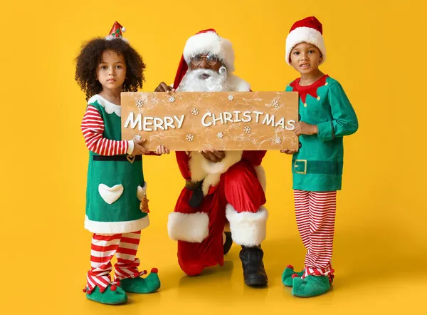 小精灵和圣诞老人拿着写着 快乐的圣诞 字样的黑板 背景是黄色的 — 图库照片