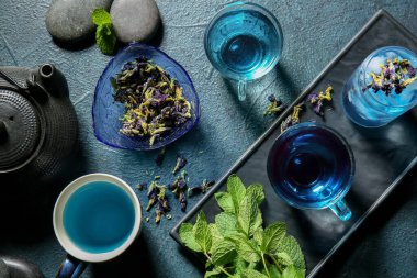 Bir bardak mavi çay, kurumuş çiçekler ve koyu arkaplanda nane.