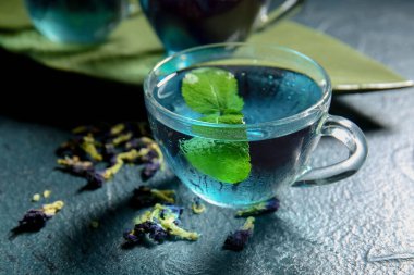 Bir bardak organik mavi çay, kurumuş çiçekler ve koyu arkaplanda nane şekeri, yakın plan.
