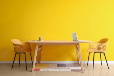 Laptoplu yemek masası, sarı duvarın yanında kahve fincanları ve dergi.