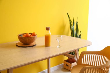 Sarı duvarın yanındaki tahta masada portakallı, meyve suyu ve cam dolu bir kase.