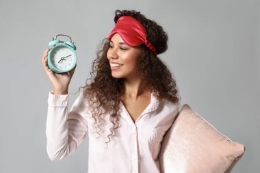 Gri arka planda uyku maskesi, çalar saati ve yastığı olan genç bir Afro-Amerikalı kadın.