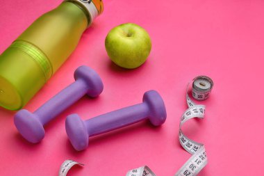 Dambıllar, mezura, pembe arka planda bir şişe su ve elma.